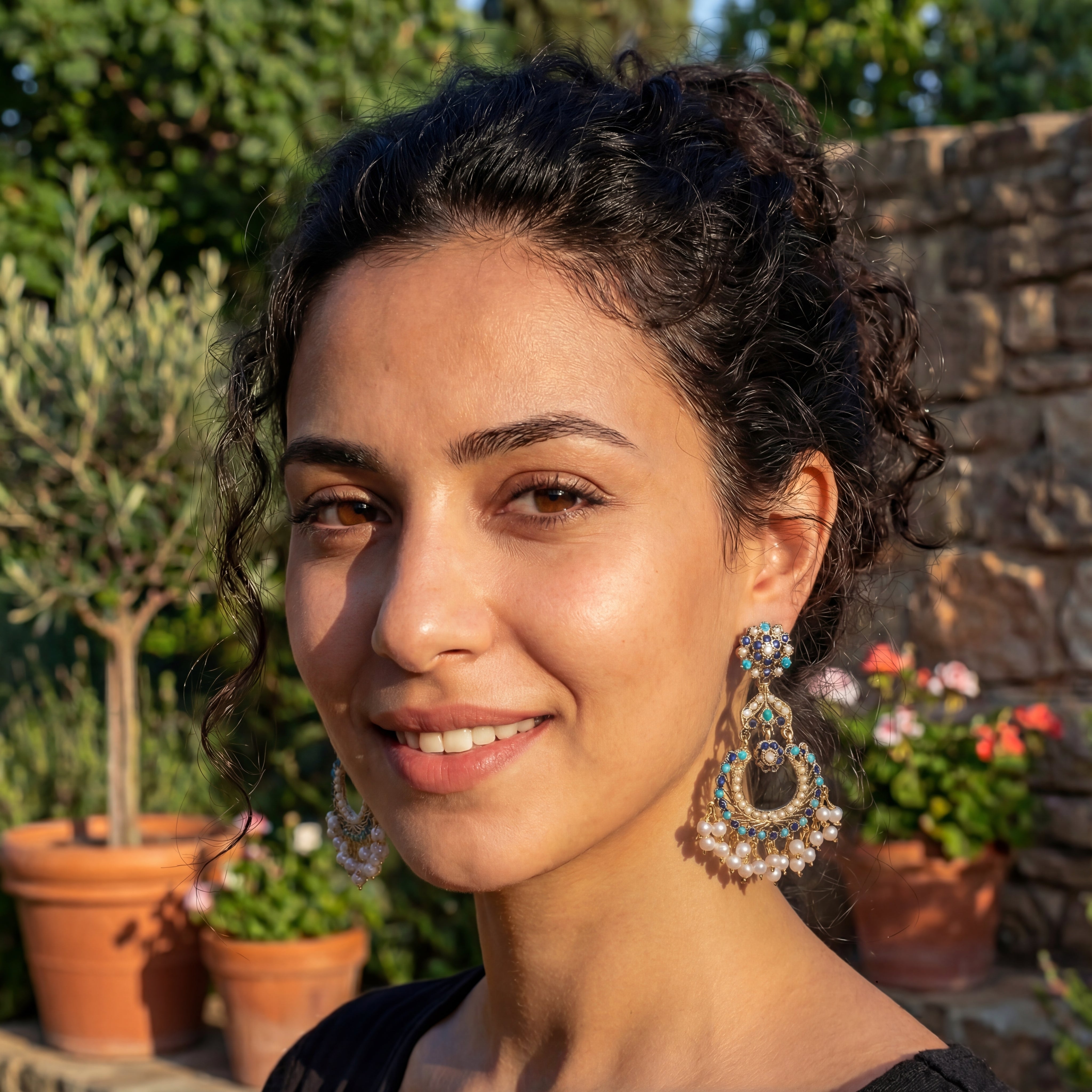 Woman with decorative earrings standing outdoors with plants and a stone wall in the background