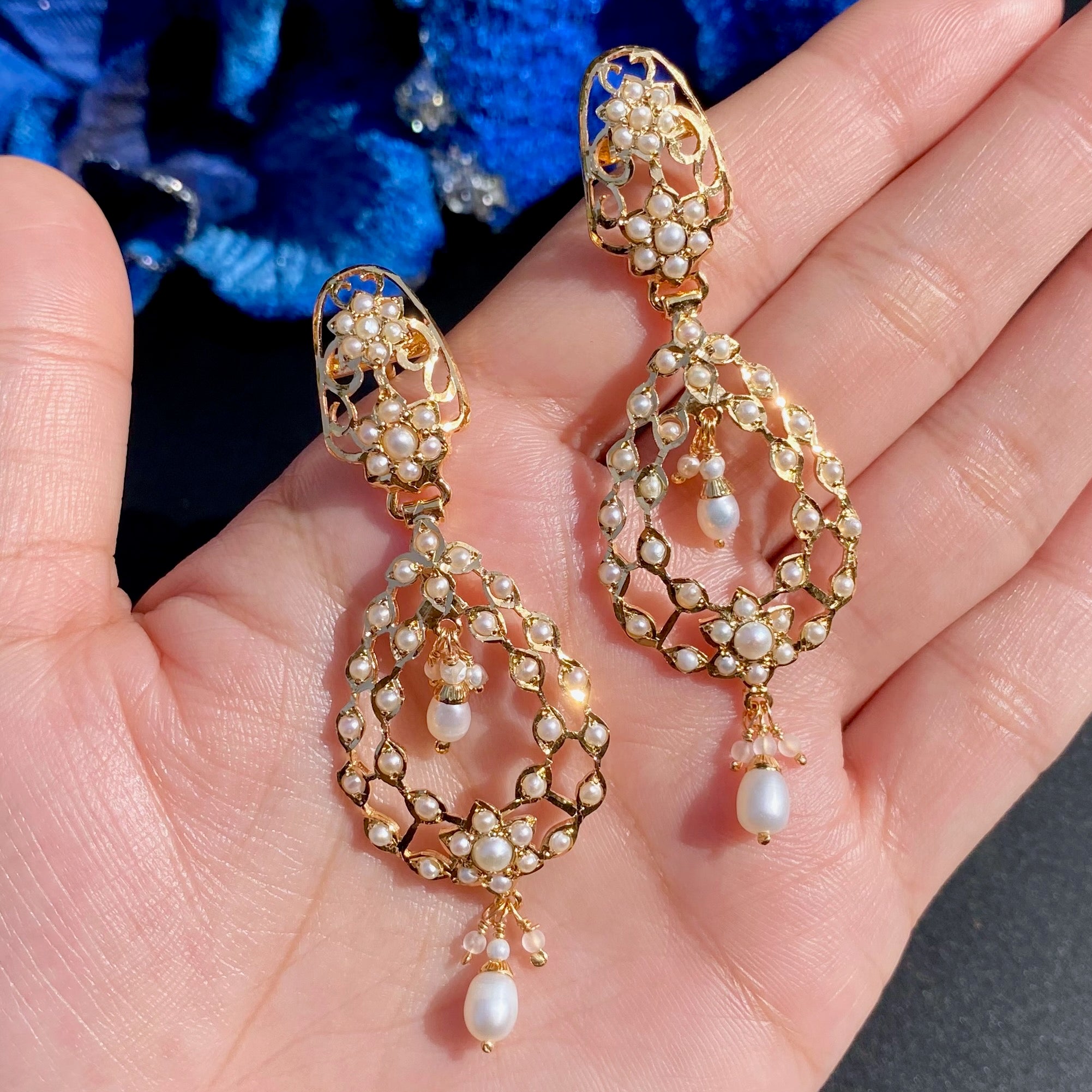 Gold earrings with pearls held in a hand against a blue floral background