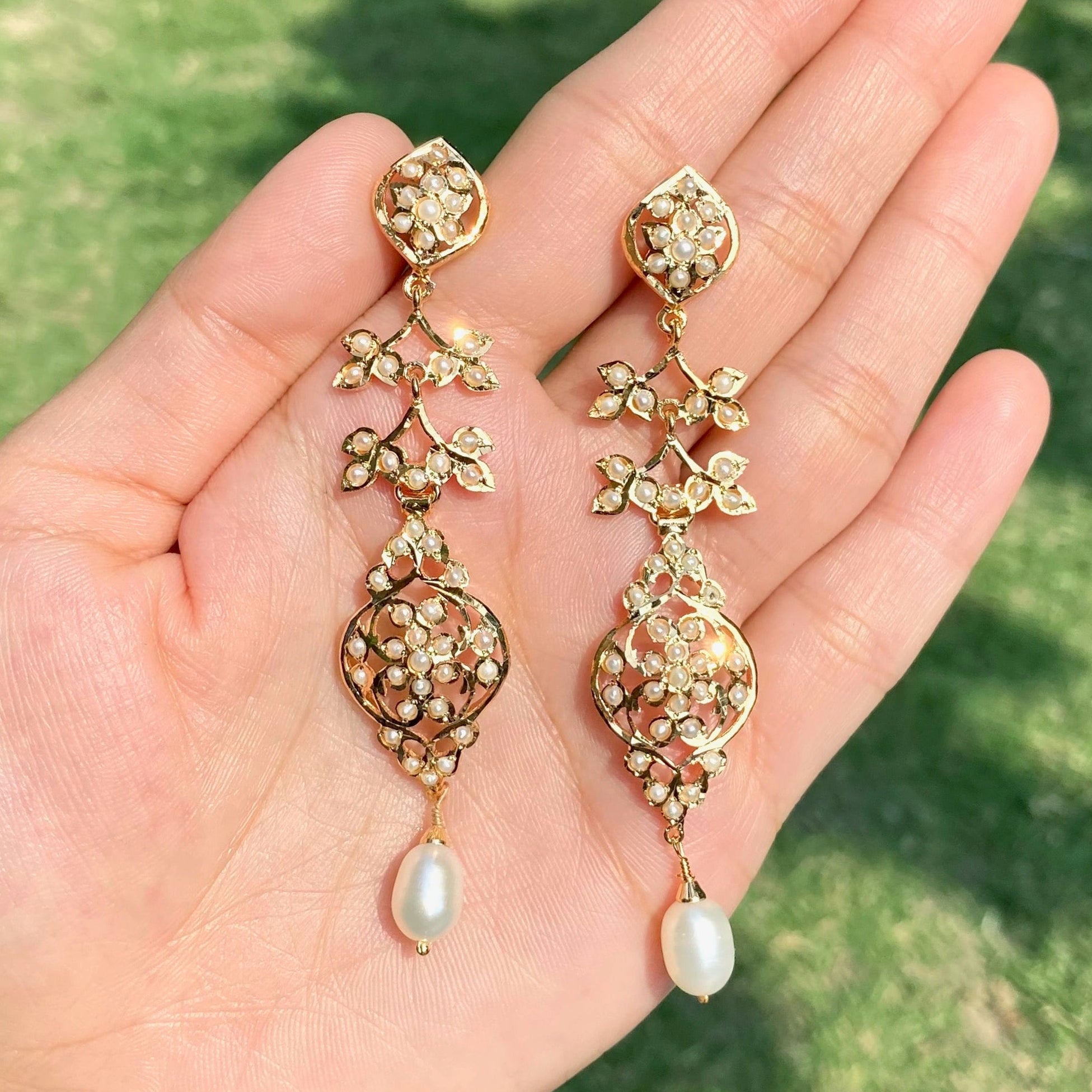 Gold earrings with pearl drops held in a hand against a grassy background