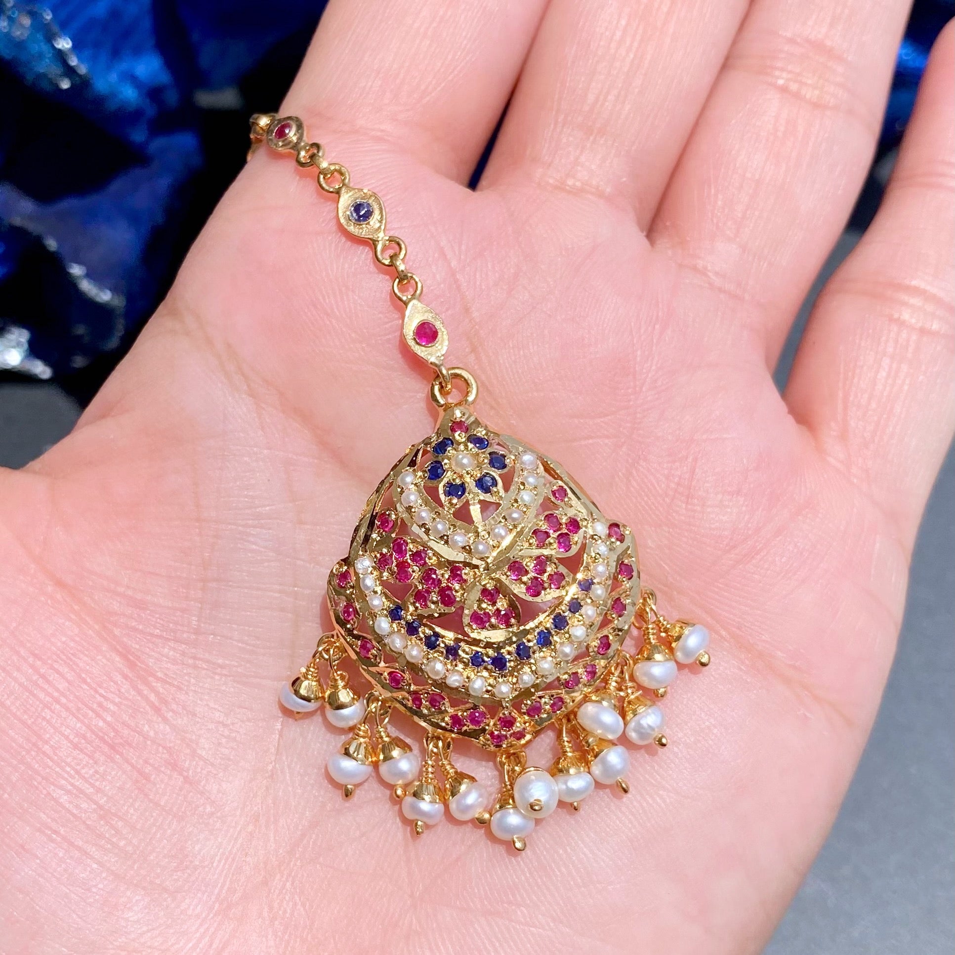 Decorative gold maang tikka with red and blue stones held in a hand against a blurred background