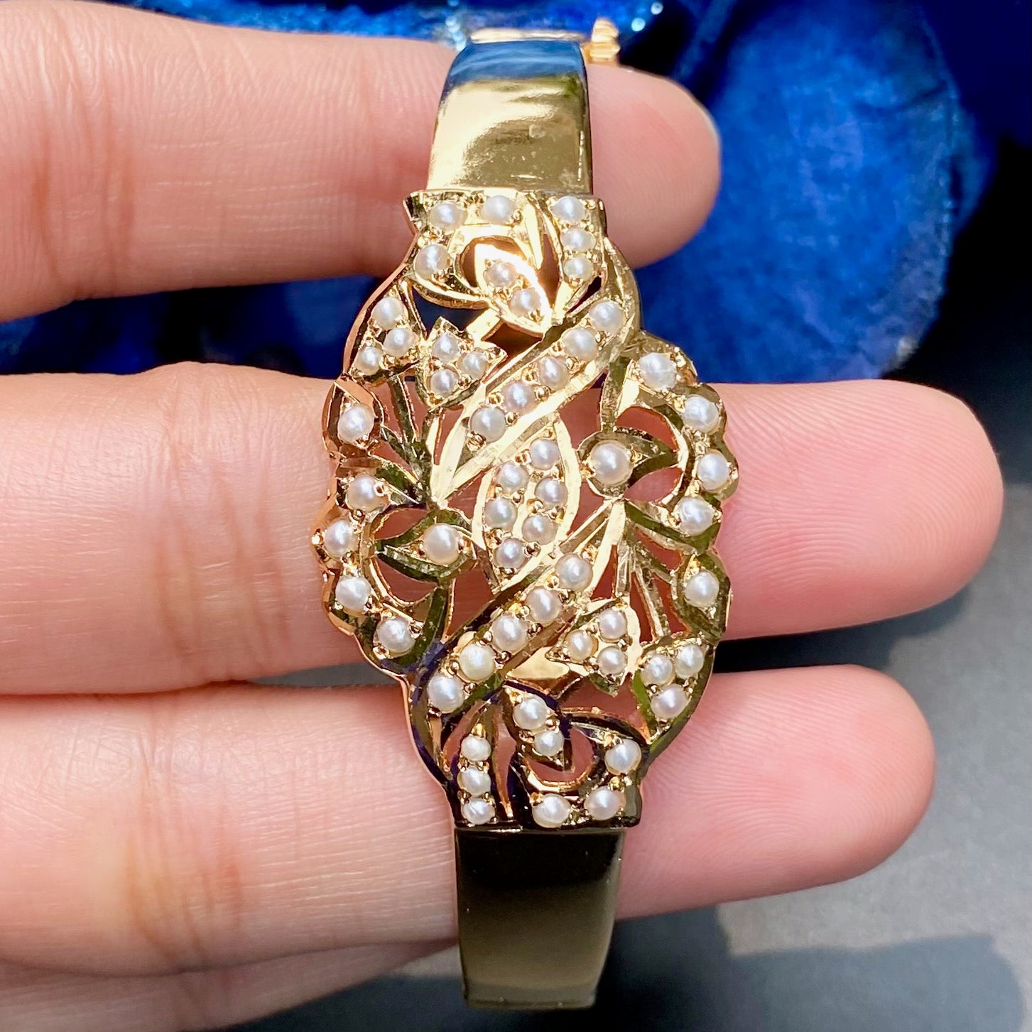 Gold bracelet with pearl accents held between fingers against a blue and gray background