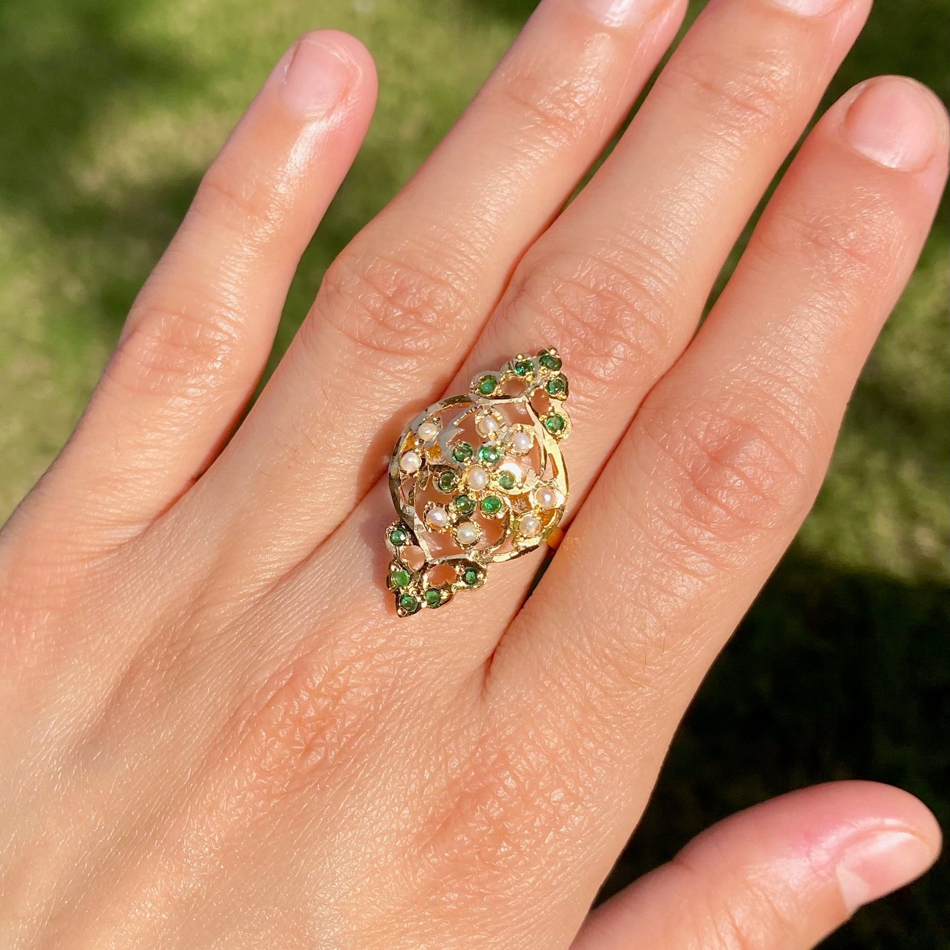 Close-up of a hand wearing a decorative ring with green gemstones against a blurred natural background