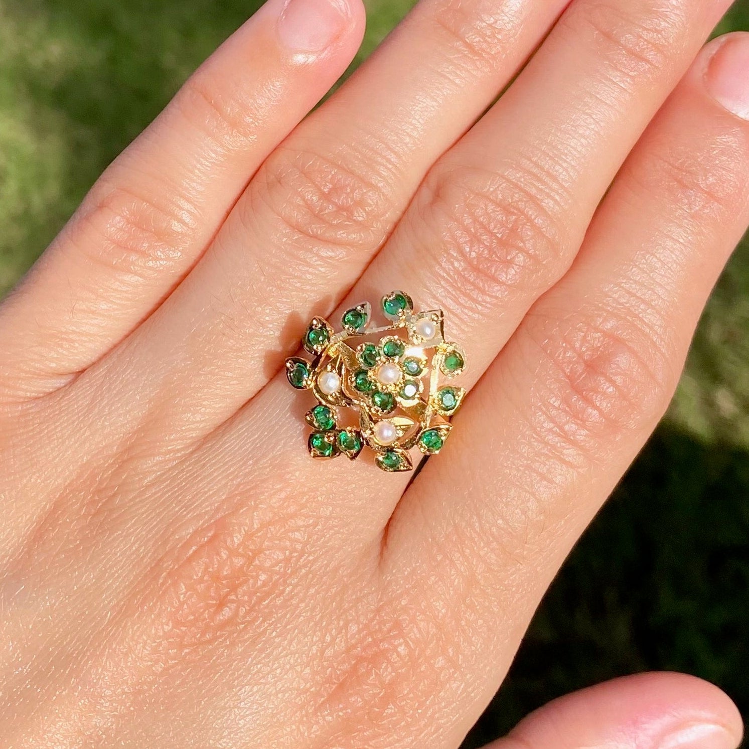 Hand wearing a ring with green gemstones against a grassy background