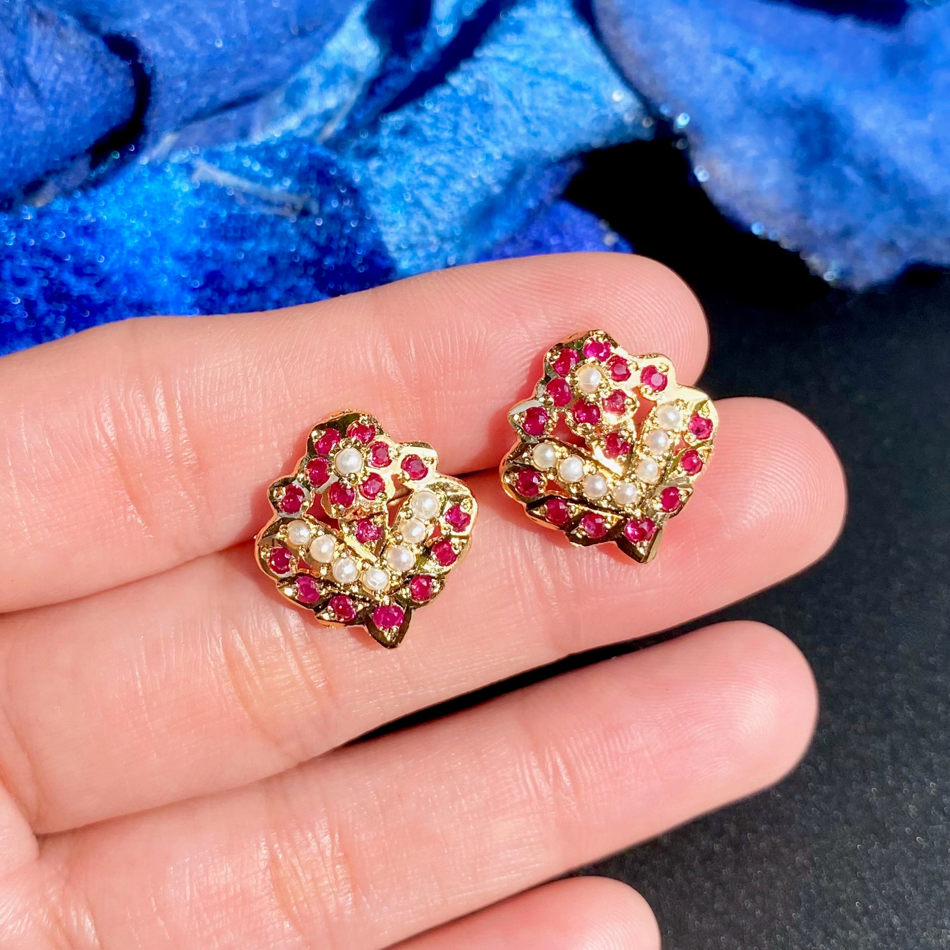 Gold earrings with red and clear gemstones held between fingers against a blue fabric background