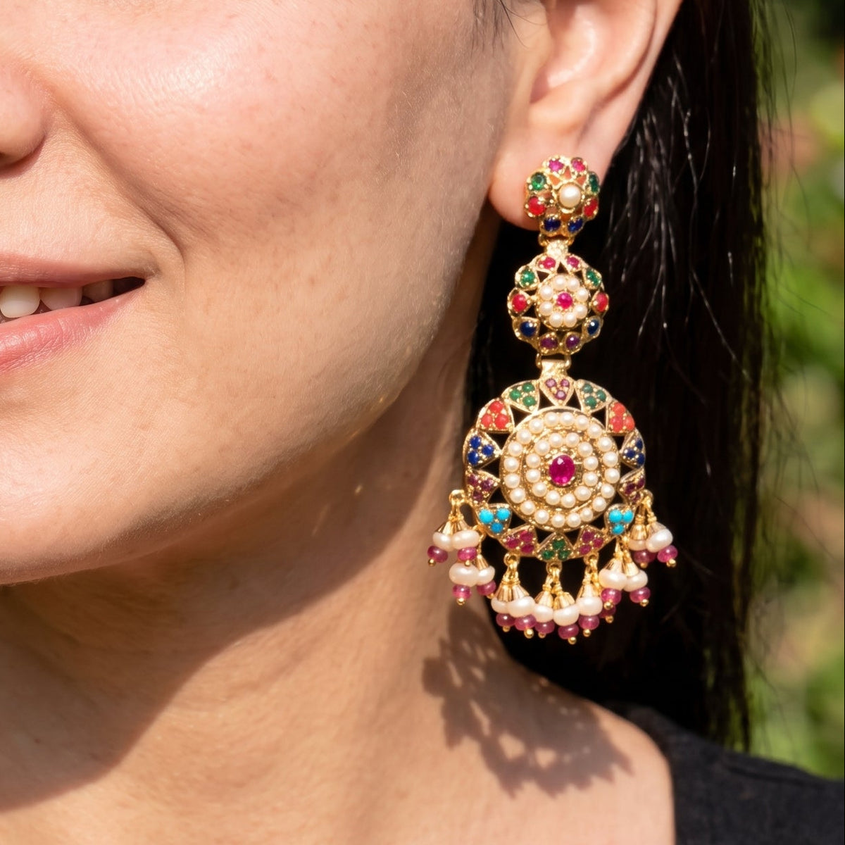 A close-up of a woman wearing ornate gold-toned chandelier earrings with colorful red, pink, and blue stone accents. The background is a softly blurred outdoor green setting.