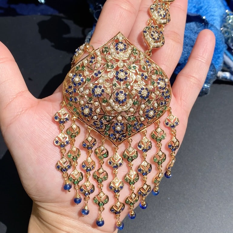 Ornate gold and gemstone necklace held in a hand against a neutral background