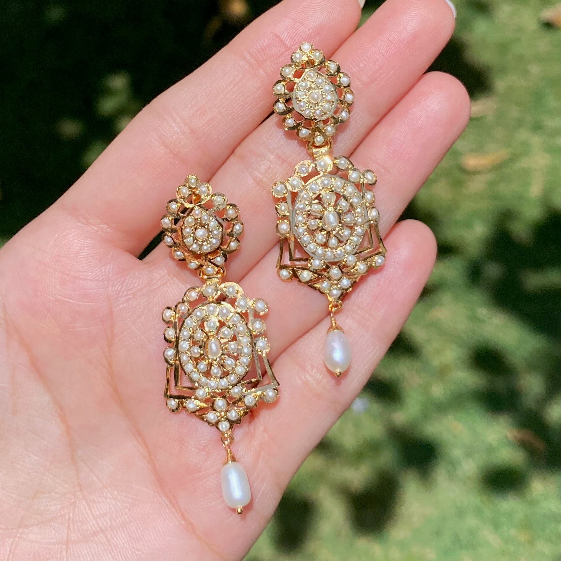 Ornate gold and pearl earrings held in a hand with a blurred green background