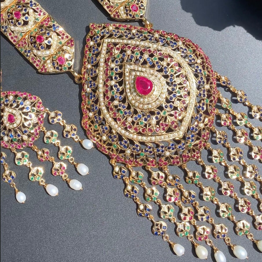 A gold-plated, long arabic-style necklace with a geometric pattern, adorned with freshwater pearls and red-blue-green stones, accompanied by a pair of matching earrings.