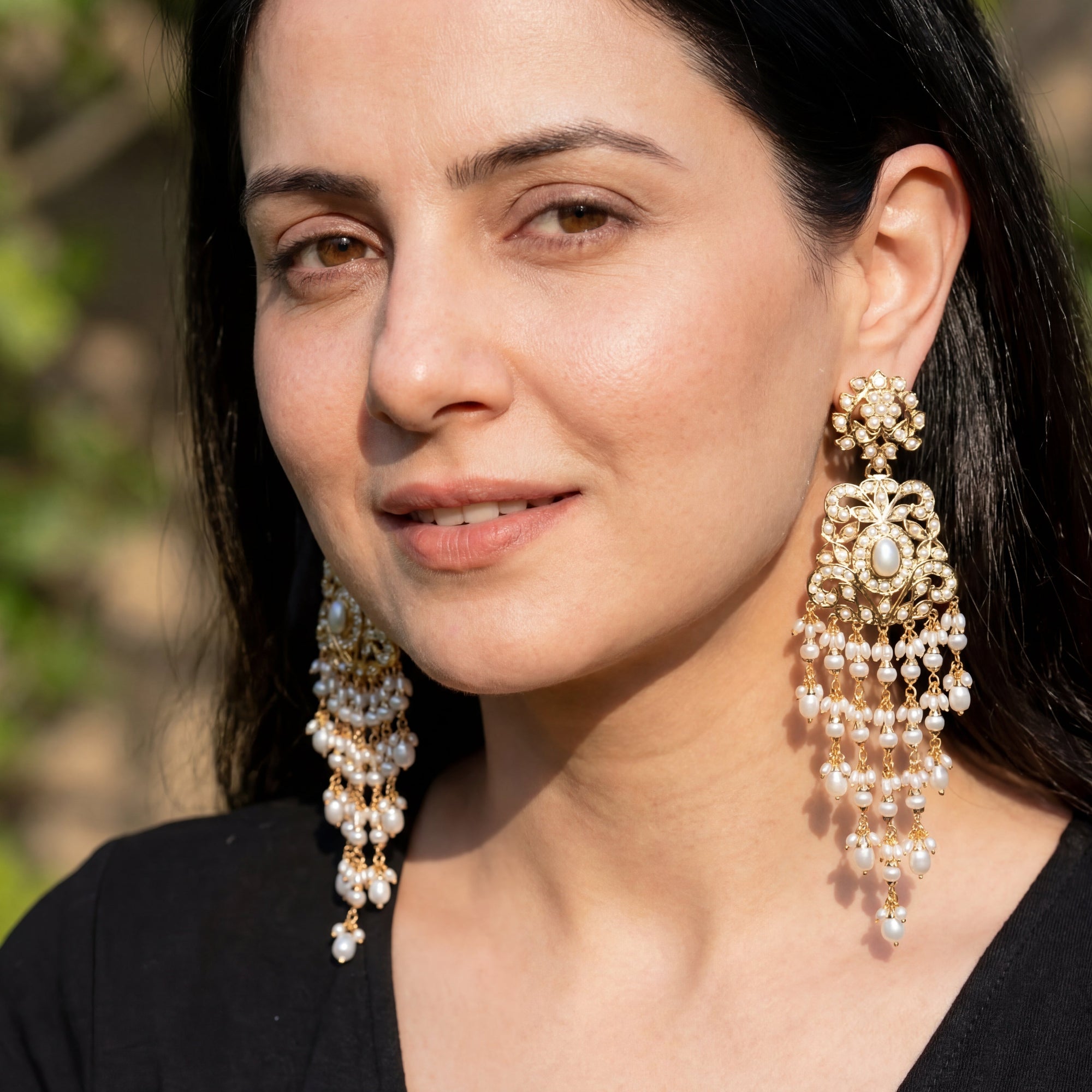 Woman wearing long gold and pearl earrings with a blurred natural background